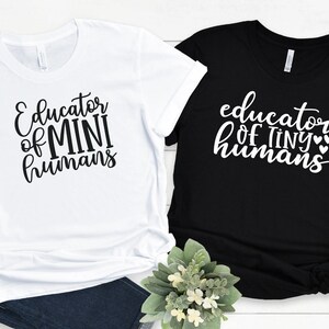 May include: Two t-shirts with a teacher theme. One white t-shirt says "Educator of Mini Humans" and one black t-shirt says "Educator of Tiny Humans" with small hearts.