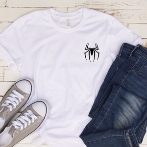 May include: White t-shirt with a black spider graphic on the front. The shirt is paired with gray sneakers and blue jeans.