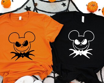 Puede incluir: Dos camisetas con un gráfico en blanco y negro de una cabeza de Mickey Mouse con la cara de Jack Skellington. La camiseta naranja tiene un gráfico negro y la camiseta negra tiene un gráfico blanco.
