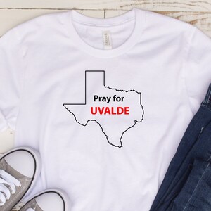 May include: White t-shirt with a black outline of the state of Texas and the text "Pray for UVALDE" in red.