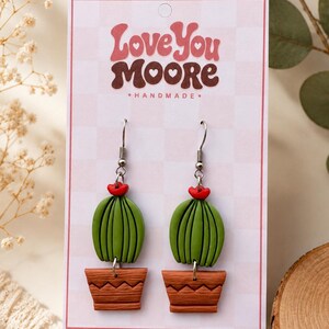May include: Handmade cactus earrings. The earrings have green cactus designs with red flower details, set in brown pot-shaped bases. The card behind the earrings reads "Love You Moore - Handmade."