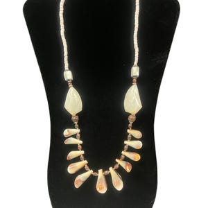 May include: A necklace with a natural, organic design. The necklace features a strand of small, white beads, with larger, teardrop-shaped pendants and shell-like elements in shades of cream, brown, and tan. The necklace is displayed on a black stand.