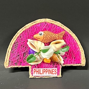 May include: A pink and brown souvenir from the Philippines featuring a fish, seashells, and flowers. The souvenir is made of woven straw and has the word "PHILIPPINES" written on it.