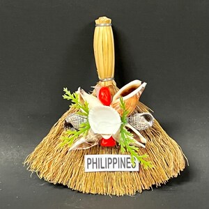 May include: A decorative miniature broom featuring a straw brush head and a woven handle. Adorned with seashells, red accents, and green sprigs. A label at the base reads "PHILIPPINES".