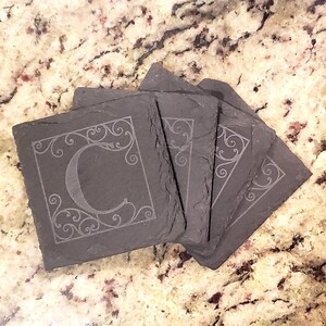 May include: Set of four black slate coasters with a decorative swirl design and the letter "C" engraved in the center of each coaster.