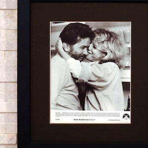 May include: Black framed print of a black and white photograph of a man and woman embracing. The print is matted in brown and the text "MAN, WOMAN AND CHILD" is visible at the bottom. The frame is hanging on a light-colored brick wall.