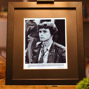 May include: Black and white framed photograph of a man with curly hair wearing a leather jacket and a collared shirt. The photograph is displayed on a wooden easel, with a gold-colored vase and a small green plant in the foreground.