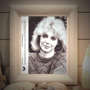 May include: A framed black and white portrait of a woman with light-colored hair, wearing a sweater and turtleneck. The frame is white with ornate detailing. Text on the side of the portrait reads "MAN, WOMAN AND CHILD".
