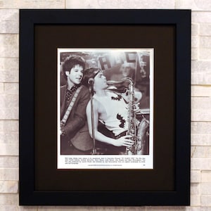 May include: Framed black and white photograph of two musicians. One plays a saxophone, the other a guitar. The image is set within a black frame and a black mount, suitable for wall display.