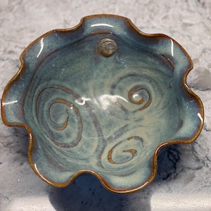 May include: A handmade ceramic bowl with a ruffled edge. The bowl features a blue-green glaze with brown spiral designs and a brown rim. A small Celtic knot design is centered on the bowl.
