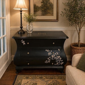May include: A black chest of drawers with three drawers and gold knobs. The chest features a curved design and floral decoration. A lamp with a beige shade sits on top, and a framed landscape painting hangs above.