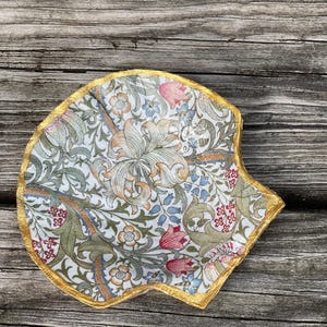 Puede incluir: Un plato decorativo en forma de concha con un estampado floral en tonos verdes, rosas y azules sobre fondo blanco. El borde es dorado. Se encuentra sobre una superficie de madera envejecida.