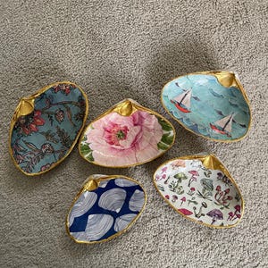 May include: Five decorative seashell dishes with gold trim. The interiors feature various designs, including floral patterns, sailboats on a blue background, and mushroom illustrations. The shells are arranged on a neutral-colored surface.
