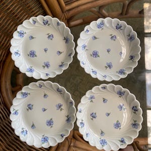 May include: A set of four white ceramic saucers with a scalloped, fluted edge design. Each saucer features a scattered pattern of small, light blue flowers with green stems and leaves printed across the center and along the curved rim.