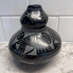 May include: A black ceramic vase with a bulbous body and a narrow neck. The vase features intricate geometric designs in a lighter color, creating a striking contrast. The handcrafted pottery showcases a traditional aesthetic.