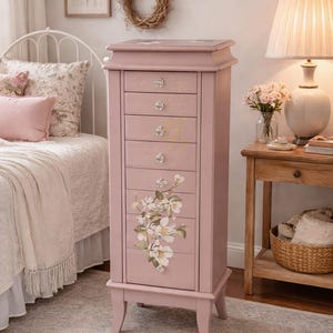 May include: A pink jewelry armoire with eight drawers and crystal knobs. The armoire features a floral design on the front and stands on four legs. The room has a bed, a lamp, and a side table.