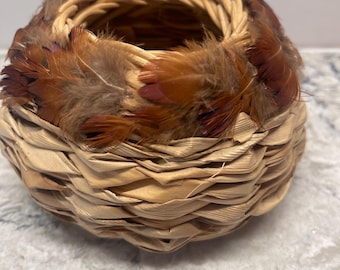 Puede incluir: Una pequeña cesta redonda tejida de color marrón claro natural. El borde está adornado con plumas marrones y rojizas. La base de la cesta está hecha de hojas secas en capas, creando una apariencia texturizada. Esta cesta decorativa es adecuada para la decoración del hogar.