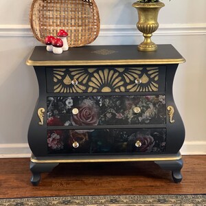 May include: A black and gold painted dresser with three drawers. The top drawer has a gold sunburst design, and the other two drawers feature a floral pattern. The dresser has gold trim and decorative hardware.