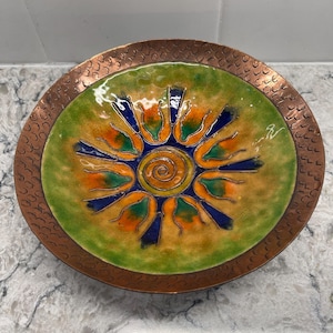 Może przedstawiać: Ozdobny miedziany talerz z kolorowym emaliowanym wzorem. Środek przedstawia wzór słońca w kolorze pomarańczowym, żółtym i niebieskim, otoczony zieloną obwódką. Obręcz ma teksturowany, młotkowany wygląd.