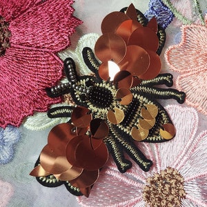 May include: Close-up of an embroidered floral design with a bee-shaped embellishment. The bee features copper-colored sequins and a black body with gold accents. The flowers are in shades of pink, peach, and blue.