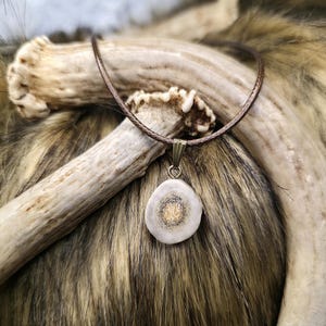May include: A pendant necklace featuring a circular, off-white pendant with a gold-colored center. The pendant hangs from a brown cord. The necklace is displayed on a backdrop of animal fur and antlers.