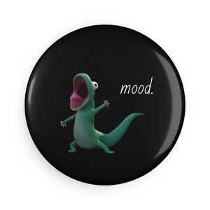 May include: A round, black button featuring a cartoon lizard with a wide open mouth and the word "mood." in white script. The lizard is green with a pink mouth and is in a dynamic pose.
