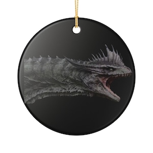 May include: A round black ceramic ornament featuring a detailed illustration of a grey dragon head with a spiky crest and an open mouth showing sharp teeth. The ornament includes a gold string loop at the top for hanging on a display hook.