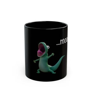 May include: Black ceramic mug featuring a cartoon lizard with an open mouth and outstretched arms. The lizard is green with a pink mouth. The text "...moo" is printed in white on the mug.