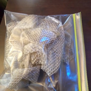 May include: A clear plastic bag containing several pieces of shed snake skin. The snake skin is a light tan color with a scale pattern. The bag has a green and yellow zipper closure.