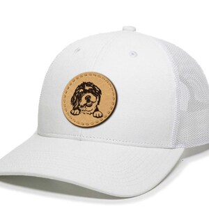 May include: White trucker hat with a mesh back and a curved brim. A round, tan leather patch features a black and brown dog illustration. The hat has a snapback closure and a structured front panel.
