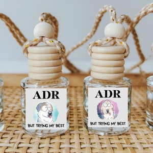 May include: Four glass car air fresheners with a wooden top and a string for hanging. Each air freshener has a cartoon dog on the front with the text "ADR BUT TRYING MY BEST".