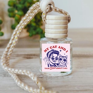 May include: A pink car air freshener with a white wooden cap and a string for hanging. The label features a cartoon woman and cat wearing sunglasses with the text "My Cat and I Talk Shit About You".