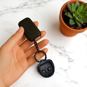 May include: Black car key fob with a matching black keychain and a decorative charm. The key fob has two buttons and is connected to a black chain and a round key ring. A small succulent plant in a terracotta pot is in the background.