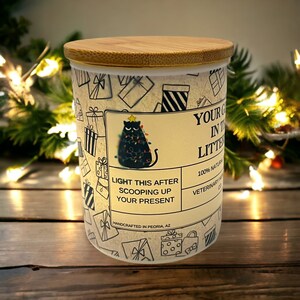 May include: A white ceramic candle jar with a bamboo lid. The jar features a black cat illustration on a Christmas tree with lights. The text on the jar reads "LIGHT THIS AFTER SCOOPING UP YOUR PRESENT" and "YOUR G IN THE LITTE 100% NATURAL VETERINARY HANDCRAFTED IN PEORIA, AZ".