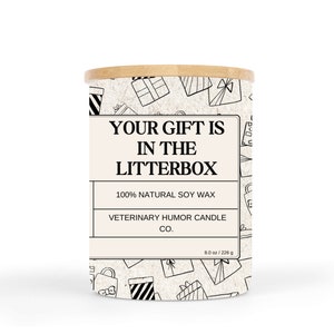 May include: A white candle jar with a black and white pattern of gift boxes. The jar has a wooden lid and a label that reads "Your gift is in the litterbox". The label also says "100% Natural Soy Wax" and "Veterinary Humor Candle Co.". The candle is 8 ounces or 226 grams.