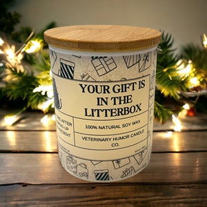May include: A white candle jar with a bamboo lid. The jar has a brown and white pattern of gift boxes and the text "Your gift is in the litterbox". The text "100% Natural Soy Wax" and "Veterinary Humor Candle Co." is also on the jar.