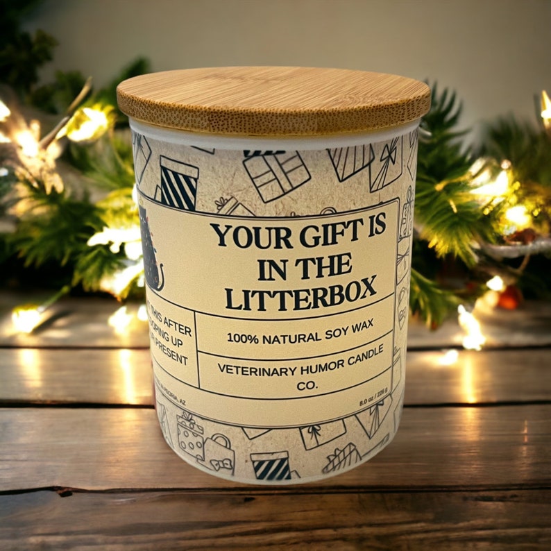 May include: A white candle jar with a bamboo lid. The jar has a brown and white pattern of gift boxes and the text "Your gift is in the litterbox". The text "100% Natural Soy Wax" and "Veterinary Humor Candle Co." is also on the jar.