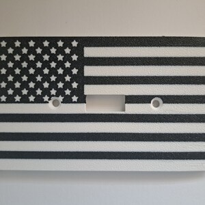 Könnte beinhalten: Ein weißer Lichtschalter-Abdeckung mit einem schwarz-weißen amerikanischen Flaggen-Design. Die Flagge hat weiße Sterne auf einem schwarzen Feld und weiße Streifen auf einem schwarzen Hintergrund.