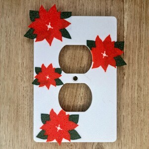 May include: White plastic outlet cover with four red poinsettia flowers with green leaves, a Christmas decoration.