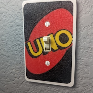 May include: A white light switch cover with a red, yellow, and black UNO card design. The switch is in the "ON" position.