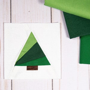May include: A white fabric square with a green and brown patchwork Christmas tree design. The fabric squares used to create the tree are shown to the right of the square.