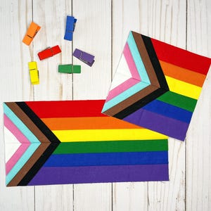 Op de afbeelding: Twee stoffen stukken met een regenboogontwerp, inclusief zwarte, bruine, roze, lichtblauwe en witte driehoeken. Zes kleine houten wasknijpers in oranje, blauw, rood, paars, geel en groen zijn er boven verspreid.