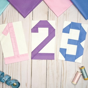 May include: Three fabric squares with the numbers 1, 2, and 3 sewn on them. The squares are pink, purple, and blue. There is a measuring tape and two spools of thread on a white wooden surface.