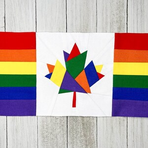 May include: A rectangular fabric quilt block featuring a central white square with a multi-colored maple leaf design composed of red, orange, yellow, green, blue, and purple segments, flanked by vertical rainbow-striped panels on both sides.
