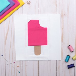 May include: A white fabric square with a pink popsicle applique. The popsicle has a brown stick and a white background. There are sewing pins and spools of thread on a wooden surface.