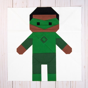 May include: A geometric quilt block featuring a stylized figure with a green body, brown arms and hands, and a black head. The figure has a green mask and a diamond shape on the chest. The background is white.