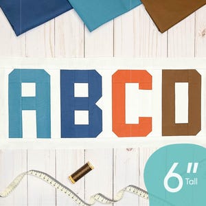Op de afbeelding: Een wit stoffen paneel met de letters ABCD in blauwe, donkerblauwe, oranje en bruine stof. De letters zijn 15,24 cm hoog. Het stoffen paneel is omgeven door restjes stof in verschillende kleuren.