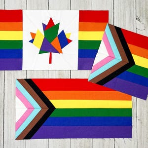 May include: Three fabric quilt blocks featuring pride-themed designs. One block displays a Canadian maple leaf composed of multicolored geometric shapes on a white background, flanked by rainbow stripes. Two blocks feature the Progress Pride flag design.
