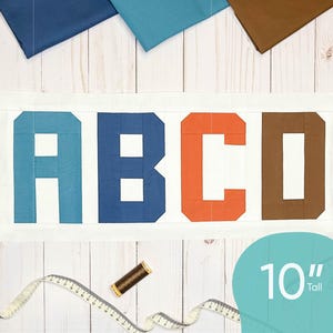 May include: A white fabric panel with the letters ABCD in blue, dark blue, orange, and brown fabric. The letters are 10 inches tall. The panel is surrounded by fabric scraps in various colors.