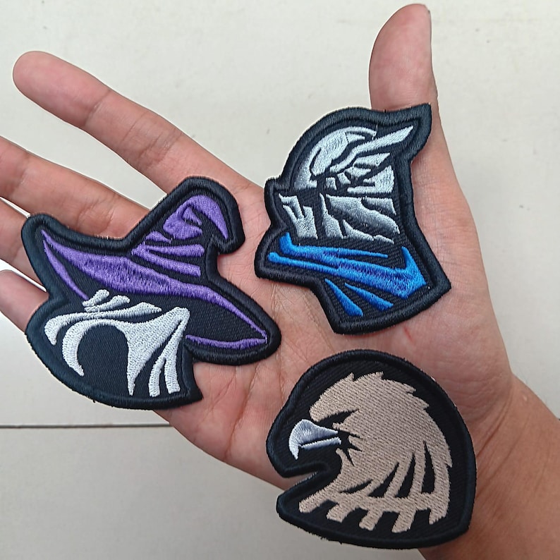 Op de afbeelding: Drie geborduurde patches: een paarse heksenhoed, een ridderhelm met blauwe accenten en een bruine adelaarskop. Elke patch heeft een zwarte rand en is ontworpen om op stof te naaien of te strijken.